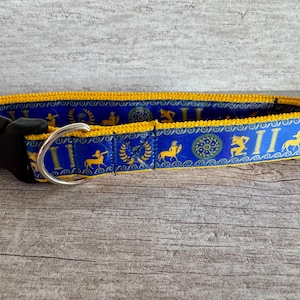 Könnte beinhalten: Ein blau-goldenes Hundehalsband mit einer schwarzen Kunststoffschnalle und einem silbernen Metall-D-Ring. Das Halsband hat ein sich wiederholendes Muster aus römischen Ziffern, Bogenschützen und Pferden. Das Halsband ist etwa 2,5 cm breit.