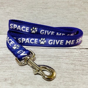 May include: A blue dog leash with a silver clip. The leash has white text that reads "SPACE GIVE ME SPACE" with a paw print between the words.