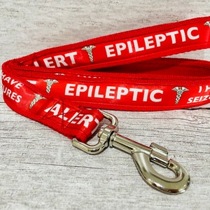 May include: Red dog leash with white text that reads "EPILEPTIC ALERT" and "I HAVE SEIZURES". The leash has a silver metal clasp and a medical symbol. The leash is designed to alert others to a dog's medical condition.