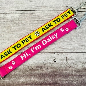 Könnte beinhalten: Zwei Hundeleinen mit Metallclips. Eine Leine ist gelb mit rotem Besatz und trägt die Aufschrift "Ask to Pet" mit einem Pfotenabdruck. Die andere Leine ist pink und trägt die Aufschrift "Hi, I'm Daisy" mit einem Pfotenabdruck.
