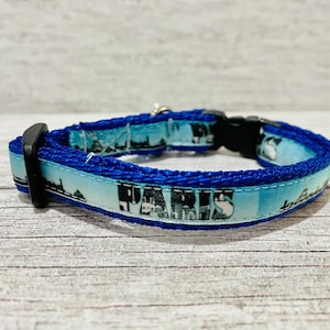 May include: Blue pet collar with a cityscape design. The collar features a light blue band with a skyline print and the word "PARIS" in black. The edges are trimmed with a darker blue fabric. It has a black plastic buckle.
