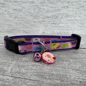 May include: A pink cat collar with a purple trim and a black buckle. The collar features a pattern of colorful donuts. A pink bell and a donut-shaped charm dangle from the collar.