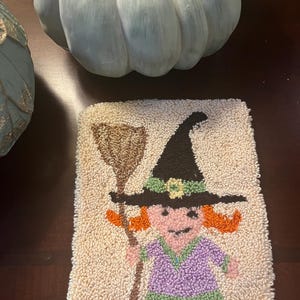 May include: A beige, rectangular, tufted rug featuring a cartoon witch with orange hair, a black hat, and a purple dress. The witch is holding a brown broom. The rug is likely handmade and suitable for Halloween decor.