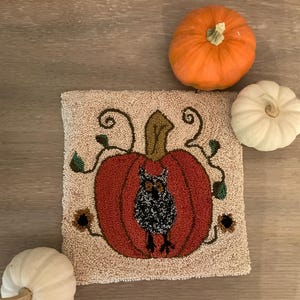 May include: A beige hand-stitched canvas with a red pumpkin and a black owl inside. The pumpkin has green vines and brown leaves. There are three white pumpkins on a wooden surface around the canvas.