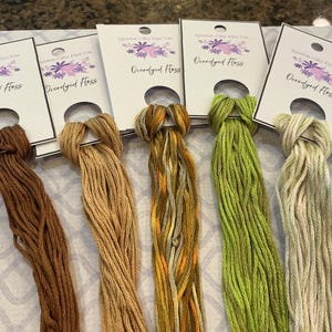 May include: Five skeins of hand-dyed embroidery floss in shades of brown, tan, yellow, green, and cream. Each skein is attached to a white card with a floral design and the text "Queen City Dye Co. Overdyed Floss."