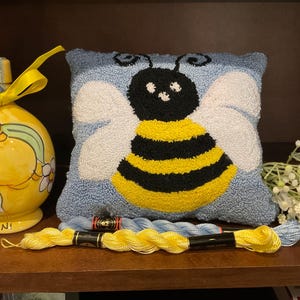 Pode incluir: Uma almofada azul e branca com um desenho de abelha amarela e preta. A almofada é cercada por fio de bordado amarelo e azul.