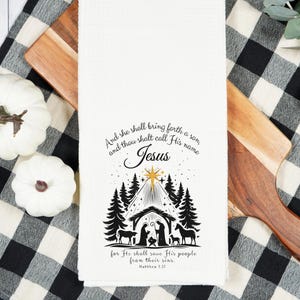 May include: White kitchen towel with a black and white Nativity scene design and the text "And she shall bring forth a son, and thou shalt call His name Jesus". The towel is on a wooden cutting board and a black and white checkered cloth.