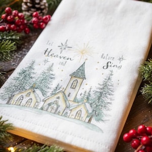 May include: White Christmas-themed kitchen towel with a church and pine tree illustration. The text "Let Heaven and Nature Sing" is printed on the towel. The towel is surrounded by pine branches, red berries, and fairy lights.