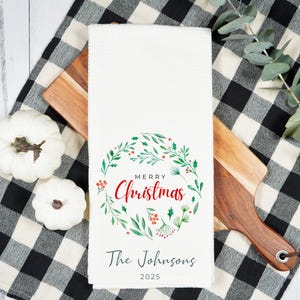 May include: White kitchen towel with a "Merry Christmas" wreath design in red and green. The towel includes the text "The Johnsons" and the year "2025". The towel is on a wooden cutting board and a black and white checkered cloth.