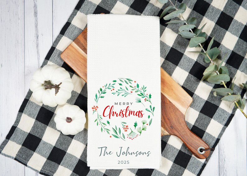 May include: White kitchen towel with a "Merry Christmas" wreath design in red and green. The towel includes the text "The Johnsons" and the year "2025". The towel is on a wooden cutting board and a black and white checkered cloth.