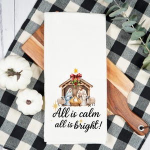May include: White kitchen towel with a watercolor Nativity scene and the text "All is calm all is bright!". The towel is placed on a wooden cutting board and a black and white checkered cloth.