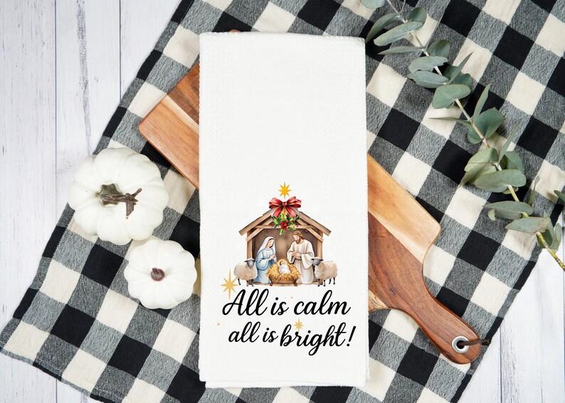 May include: White kitchen towel with a watercolor Nativity scene and the text "All is calm all is bright!". The towel is placed on a wooden cutting board and a black and white checkered cloth.