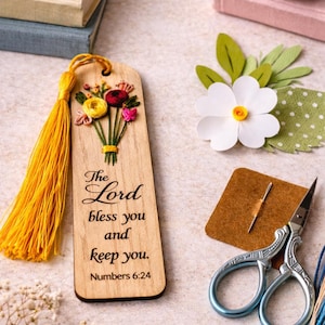May include: Wooden bookmark with embroidered flowers and a yellow tassel. The text reads "The Lord bless you and keep you. Numbers 6:24." A white paper flower and small scissors are also visible.