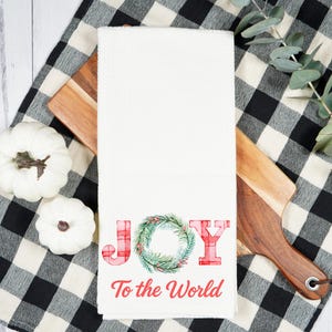 May include: White waffle-weave kitchen towel with the words "JOY To the World" in red and a green wreath design. The towel is on a wooden cutting board, with white pumpkins and a black and white checkered cloth.