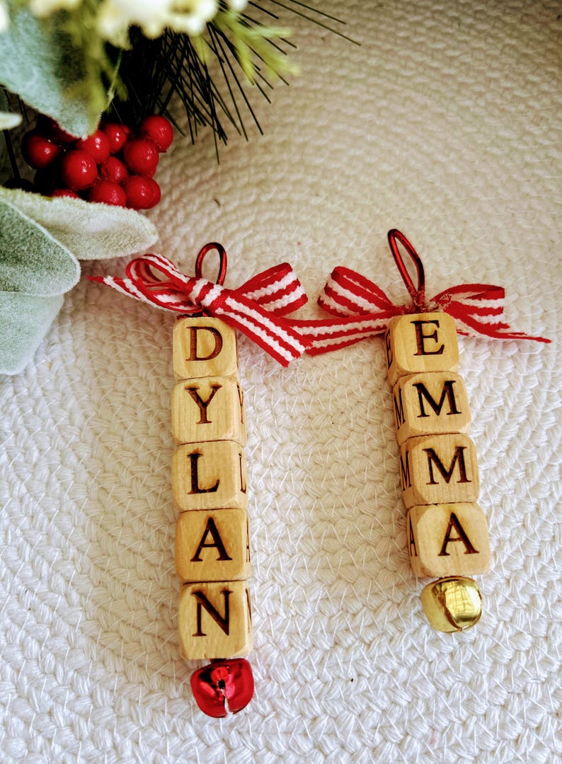 以下が含まれることがあります： 「Dylan」と「Emma」と書かれた木製の文字ブロックを使った、2つのパーソナライズされたクリスマスオーナメント。各オーナメントには、赤と白のストライプのリボンと、下部に小さなベルが付いています。お祝いのホリデーデコレーション。