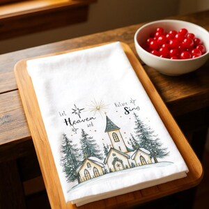 May include: White kitchen towel with a church scene and the text "Let Heaven and Nature Sing." The design includes a church, pine trees, and a starburst. The towel is on a wooden tray.