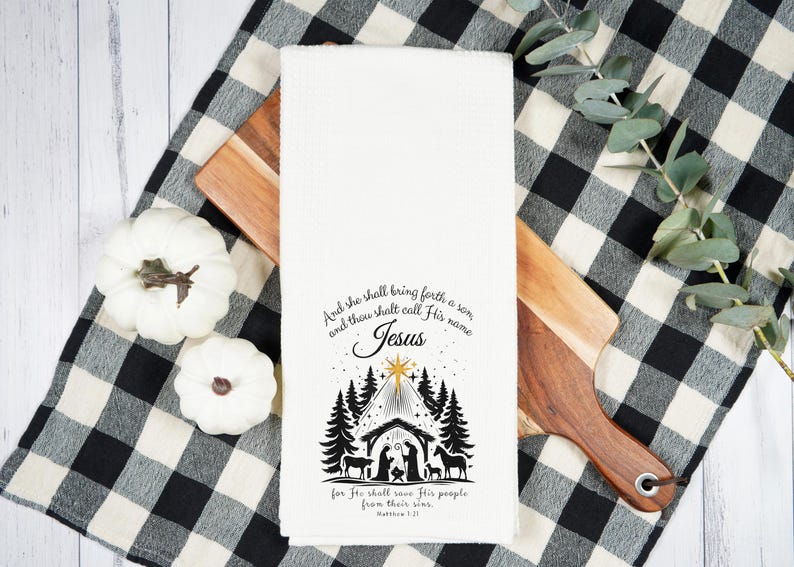 May include: White kitchen towel with a black and white Nativity scene design and the text "And she shall bring forth a son, and thou shalt call His name Jesus". The towel is on a wooden cutting board and a black and white checkered cloth.