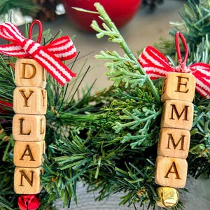 以下が含まれることがあります： 「DYLAN」と「EMMA」と書かれた木製の名前飾り。赤と白のストライプのリボンと小さなベルが付いています。緑の松葉やその他の緑の葉の上に飾られており、背景には赤い飾りがあります。