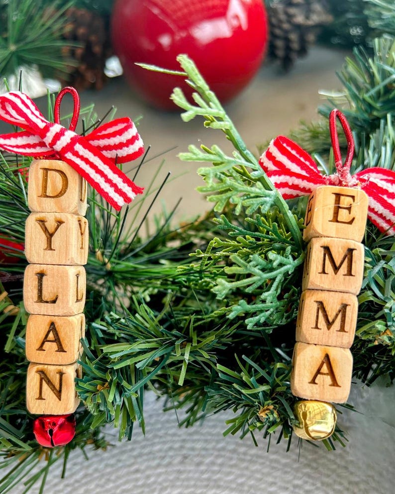 以下が含まれることがあります： 「DYLAN」と「EMMA」と書かれた木製の名前飾り。赤と白のストライプのリボンと小さなベルが付いています。緑の松葉やその他の緑の葉の上に飾られており、背景には赤い飾りがあります。