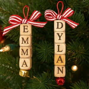 以下が含まれることがあります： 「EMMA」と「DYLAN」と書かれた2つのパーソナライズされたクリスマスオーナメント。各オーナメントは、文字が書かれた木製のブロックでできており、赤と白のストライプのリボンで飾られ、小さなベルで仕上げられています。オーナメントは、緑の松の葉を背景に吊り下げられています。