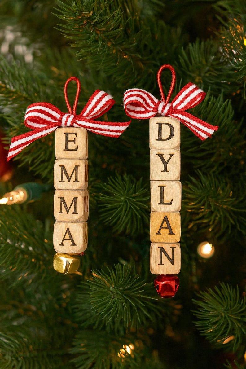 以下が含まれることがあります： 「EMMA」と「DYLAN」と書かれた2つのパーソナライズされたクリスマスオーナメント。各オーナメントは、文字が書かれた木製のブロックでできており、赤と白のストライプのリボンで飾られ、小さなベルで仕上げられています。オーナメントは、緑の松の葉を背景に吊り下げられています。
