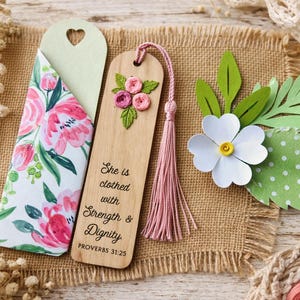 May include: Wooden bookmark with embroidered flowers and a pink tassel, alongside a floral print bookmark holder. The wooden bookmark has the text "She is clothed with Strength & Dignity" from Proverbs 31:25. A paper flower and leaves are also present.