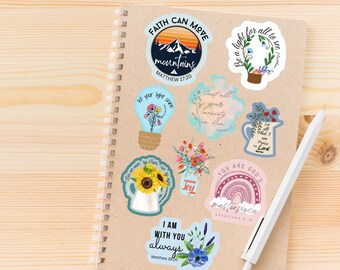 May include: A notebook with colorful stickers featuring inspirational quotes and bible verses. The stickers include images of a mountain, a lightbulb, a sunflower, a rainbow, and a jar of flowers. The notebook is on a wooden surface.