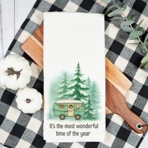 May include: White kitchen towel with a watercolor illustration of a teal camper in front of green pine trees. The text "It's the most wonderful time of the year" is printed at the bottom. The towel is on a wooden cutting board and a black and white checkered cloth.