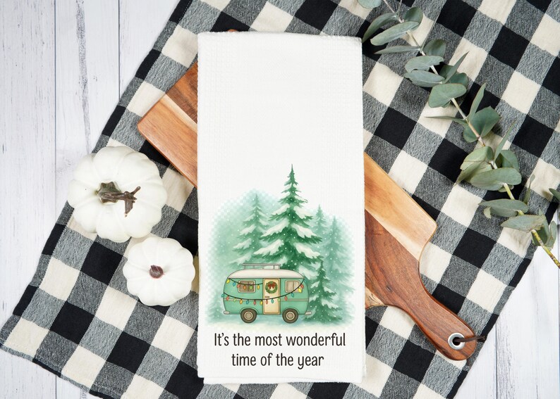 May include: White kitchen towel with a watercolor illustration of a teal camper in front of green pine trees. The text "It's the most wonderful time of the year" is printed at the bottom. The towel is on a wooden cutting board and a black and white checkered cloth.