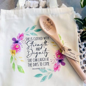 May include: A white tote bag with a floral design and the quote "She is clothed with strength and dignity. She can laugh at the days to come. Proverbs 31:25". A wooden spoon with the quote "Be on your guard, stand firm in the faith, be courageous, be strong." is included. A small wooden tag with the text "Happy Mother's Day" is attached to the spoon.