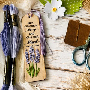May include: A wooden bookmark with embroidered lavender flowers and the text "HER CHILDREN rise up AND CALL HER blessed Proverbs 31:28". The bookmark has a purple tassel and is next to spools of blue embroidery floss.