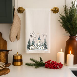 May include: A white kitchen towel with a winter scene featuring a church, evergreen trees, and the text "Let Heaven and Nature Sing." The towel is hanging on a brass towel rack, with candles and greenery nearby.