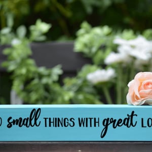 May include: A light blue wooden sign with the text "Do small things with great love" in black cursive lettering. The sign is sitting on a wooden surface with a pink rose and white flowers in the background.
