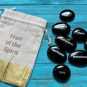 May include: A set of black stones with words representing the fruit of the spirit, including love, peace, joy, gentleness, goodness, self-control, faith, kindness, and forbearance. The stones are in a small, beige drawstring bag with the words "Fruit of the Spirit" printed on it.