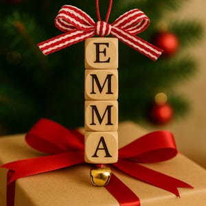 以下が含まれることがあります： 赤いリボンと「EMMA」と書かれた木製オーナメントが付いたクリスマスをテーマにしたギフトボックス。オーナメントは、E、M、M、Aの文字が入った4つの木製ブロックでできています。上部には赤と白のストライプのリボンがあり、下部には小さな金のベルが付いています。