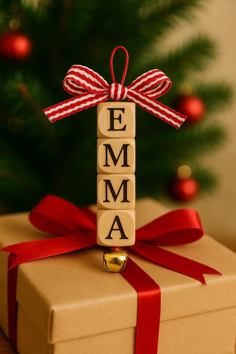 以下が含まれることがあります： 赤いリボンと「EMMA」と書かれた木製オーナメントが付いたクリスマスをテーマにしたギフトボックス。オーナメントは、E、M、M、Aの文字が入った4つの木製ブロックでできています。上部には赤と白のストライプのリボンがあり、下部には小さな金のベルが付いています。