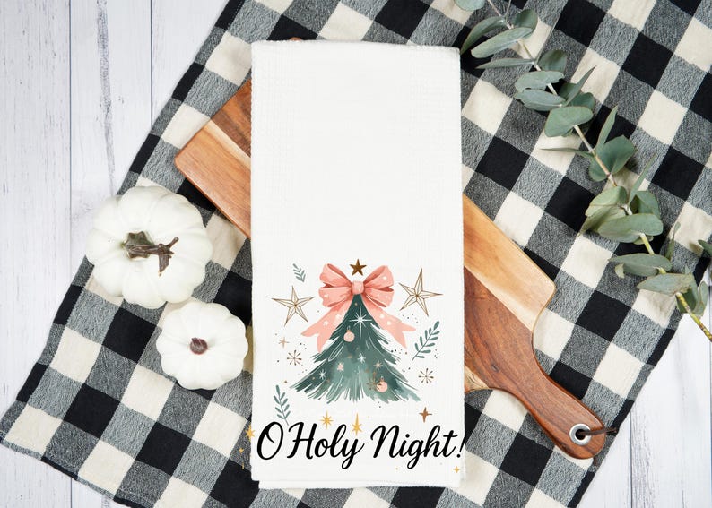 May include: A white kitchen towel with a watercolor Christmas tree design, a pink bow, and the text "O Holy Night!" is displayed on a wooden cutting board and a black and white checkered cloth. Decorative pumpkins are also present.