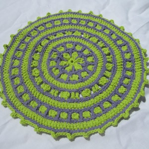 May include: A round crocheted doily with a purple and green design. The doily has a scalloped edge.