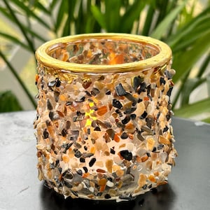 May include: A decorative glass candle holder with a gold rim, encrusted with small, colorful stones. The stones are shades of brown, black, and beige. The candle holder is lit, casting a warm glow.