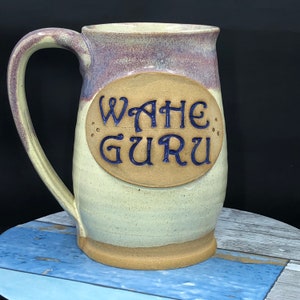 May include: A ceramic mug with a light brown glaze and a purple and white glaze on the top. The mug has a large oval-shaped label with the text "WAHE GURU" in blue lettering.