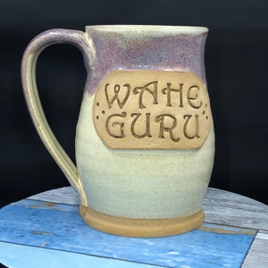 May include: A ceramic mug with a light brown glaze and a purple rim. The mug has a handle and a large inscription that reads "Wahe Guru".