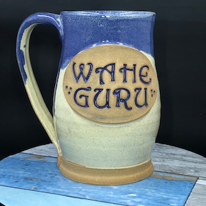 May include: A ceramic mug with a blue and white glaze. The mug has a handle and a large oval on the front with the words "Wahe Guru" in blue lettering.