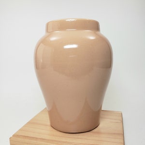 May include: A light brown ceramic vase with a smooth, glossy finish. The vase is sitting on a wooden base.
