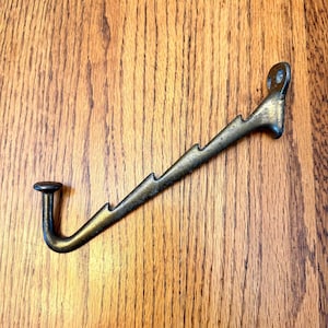 May include: A bronze-colored metal hook with a decorative, jagged design along the angled arm. The hook has a rounded end and a loop at the top for hanging. The hook is set against a wooden background.