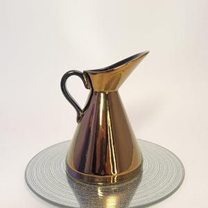 May include: A metallic gold-colored ceramic pitcher with a black handle and spout. The pitcher has a conical shape and sits on a clear, circular glass plate. The surface of the pitcher reflects light, creating a shiny appearance.
