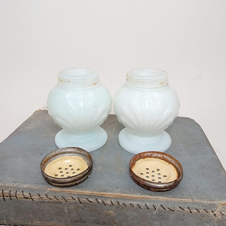 May include: Two antique, opalescent white glass salt shakers with decorative floral patterns. Each shaker has a round base and a bulbous body. The metal lids are off, revealing the interior. The shakers are displayed on a textured, gray surface.