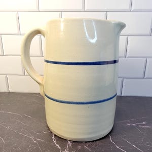 May include: Cream-colored ceramic pitcher with a large handle and spout. Two horizontal blue stripes encircle the body of the pitcher. The pitcher is a classic design, suitable for serving beverages.