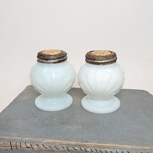 May include: A pair of antique white glass salt shakers with silver-colored metal lids. The shakers have a bulbous body with a textured, ridged design and a pedestal base. The lids have small holes for dispensing salt.