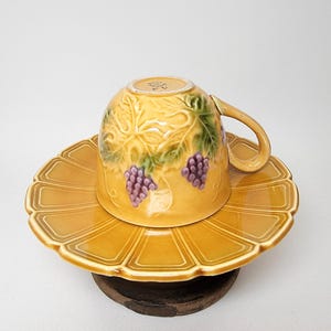 May include: A yellow ceramic teacup and saucer set with a grapevine design. The cup and saucer are decorated with green leaves and purple grapes.