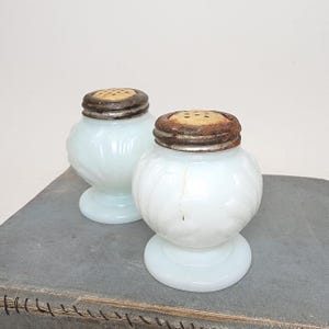 May include: Two antique, opalescent glass salt and pepper shakers. The shakers have a bulbous body with a decorative swirl pattern and sit on a round base. The lids are metal with small holes for dispensing. The shakers are displayed on a gray surface.